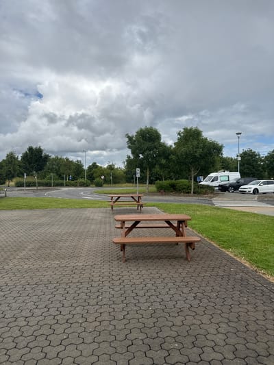 The image shows an outdoor seating area with picnic tables situated on a hexagonal paver stone ground. In the background, there is a road with white lane markings, surrounded by green grass and trees. Several vehicles, including a white van, a black car, and a white car, are parked on the side of the road. There are also streetlights and various traffic signs visible. The sky is overcast with heavy clouds. The location is identified as Commons Cross, Ireland. There are no people or any interactions depicted in the image.