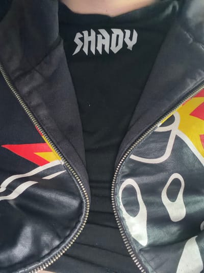 The image captures a close-up, downward-angled view of a person's upper torso and neck, located in the city of Karadjovo, Bulgaria. The individual is wearing a black t-shirt with the word "SHADY" printed in stylized, jagged white capital letters across the upper chest. Over this, a black zippered jacket or hoodie is worn, partially unzipped down the center to reveal the t-shirt beneath. The jacket features a metallic zipper. The left side of the jacket (viewer's right) displays a design with red and yellow angular patterns alongside white curved lines. The right side of the jacket (viewer's left) exhibits irregular white, abstract shapes against a black background, with this section appearing to have a distinct, possibly reflective or faux leather texture compared to the fabric of the left side. No other subjects, objects, or environmental details are discernible.
