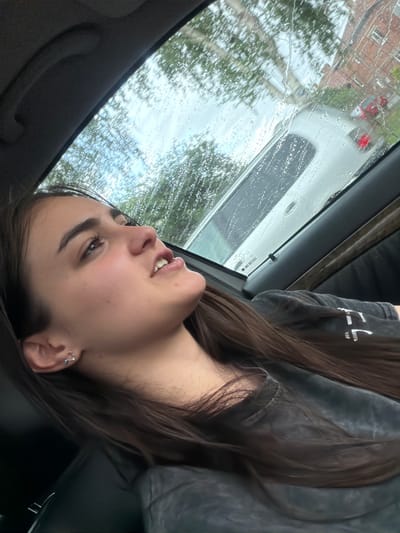 A young woman with long brown hair is reclining in the passenger seat of a car. She is wearing a grey shirt. The car window is covered in raindrops, and the blurry background shows a street with buildings and trees. A red car is visible in the background. The location appears to be urban. Barakovo, Bulgaria is not discernible in the image.