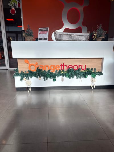 The image displays the reception area of an Orangetheory Fitness studio in Santo Domingo, Dominican Republic. A white and wood-paneled reception desk is centrally positioned, decorated for the holidays. A green garland, interspersed with small green spherical ornaments, two cream-colored dreamcatchers with feathers, and small wooden heart cutouts, hangs along the front of the desk. On the white countertop, from left to right, are a small artificial Christmas tree with red berries, a "December" calendar, and a rectangular woven basket containing rolled items, enclosed in clear plastic. Another small artificial Christmas tree is visible on the far right of the desk. The wall behind the desk is solid red and features a large, metallic silver Orangetheory logo (stylized heart-rate monitor icon). To the left, a glass entrance or partition reflects a Grinch-themed holiday decoration showing a green hand holding a red ornament. The floor consists of large, matte grey tiles with visible grout lines and subtle reflections. No individuals are present in the scene, and no actions or interactions are depicted.