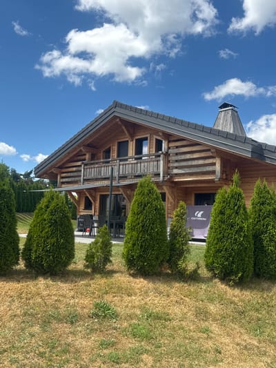 The image captures a charming wooden chalet-style house situated in a grassy area, with a clear blue sky and scattered white clouds overhead. The house features a dark grey roof and a prominent wooden chimney. In front of the house, a row of conical evergreen trees stands tall, partially obscuring the lower facade. A small black canopy extends from the house, providing shade over an outdoor seating area with black chairs and tables. On the lower right side of the house, a large purple banner with a white logo and text is visible. The foreground consists of dry, golden-brown grass with scattered green patches and small yellow wildflowers. The background reveals more lush greenery, suggesting a natural setting. The overall impression is one of a serene, possibly rural or semi-rural environment, consistent with a location like Les Rousses, France, which is known for its natural beauty and outdoor activities. No people are visible in the image.