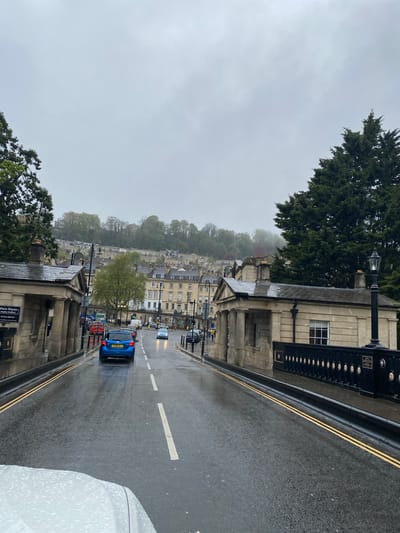 The image depicts a street scene in Bath, United Kingdom on a rainy day. The road is wet and reflects the overcast sky. Yellow double lines run along the edge of the road, and a broken white line marks the center.
A blue car is traveling down the road, away from the viewer. Other vehicles, including what appears to be a red double-decker bus, are visible further down the street.
Buildings line the street, constructed in a classic Bath architectural style, featuring light-colored stone. A tree is visible in the middle ground. Above the buildings, a hillside ascends, with more buildings and trees visible through the mist.
In the foreground on the right, the side of a white vehicle is partially visible. On the left, a stone structure with a metal railing and lamp posts runs alongside the road. A sign is visible on the structure closer to the viewer, but the text is illegible. The overall atmosphere is damp and misty.