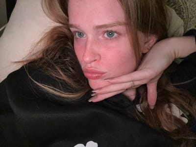 A close-up, high-angle image depicts a young adult female lying on a light-colored surface, possibly a pillow. She has fair skin, blue eyes, and long, wavy reddish-brown hair spread around her head and left shoulder. Her lips are pursed in a 'kiss' expression, and her gaze is directed upwards and slightly to her left. Her right hand is positioned with fingers extended beneath her chin, displaying light-colored nail polish and a thin, gold-toned ring on her ring finger. Her left ear is visible, adorned with a small stud and a small hoop earring. She is wearing a dark, long-sleeved garment, likely a sweatshirt, with a partial white design visible on the lower left portion. A patterned fabric, possibly part of a cushion or throw, is visible in the upper right background. The specific location in Rosmalen, Netherlands, cannot be determined from the visual information provided.