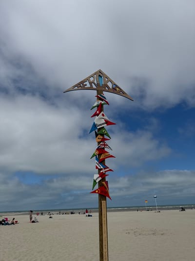 An upward-pointing arrow-shaped signpost with various national flags and greetings in different languages stands on a sandy beach. The signpost is wooden and is adorned with small triangular flags, each featuring a country's flag or a greeting such as "Bienvenue," "Welcome," and "Willkommen." At the top of the signpost, cut-out letters spell out "BERCK." In the background, a calm ocean meets the horizon under a cloudy sky. Several people are scattered across the beach, some sitting or lying on towels, while others are walking. Two flags on poles, one yellow and one blue and white, are visible further down the beach.