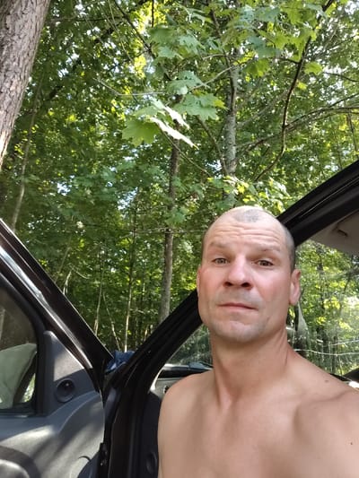 The image is a close-up, eye-level selfie taken outdoors, likely in a wooded area. The subject is a fair-skinned, middle-aged male with a shaved head and light stubble. He is shirtless and looking directly at the camera with a neutral expression. The background consists of lush green trees and foliage, with the open door of a vehicle visible on the left side of the frame. The lighting suggests it is daytime, with sunlight filtering through the leaves. There are no clear indicators of a specific urban location; the environment appears rural or natural.