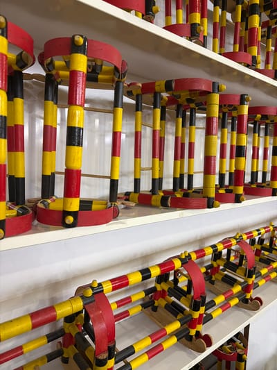 The image displays a close-up, eye-level view of multiple handmade wooden toys on white shelves. These toys are constructed from painted wooden rods, predominantly in red, yellow, and black, arranged in various configurations. The toys appear to be part of a collection or display, with multiple identical or similar units.

In the foreground, on a lower shelf, a toy constructed of red, yellow, and black rods is visible. It features a framework of horizontal and vertical rods, with curved red elements attached to the top. The bottom of this toy rests on what appear to be small wooden sled-like bases.

On the shelf above, several more of these toys are arranged. They share the same color scheme and construction style, with vertical posts connected by horizontal rods and capped with red curved pieces. The overall impression is one of a playful and colorful exhibit.

The background consists of plain white walls and shelves, suggesting an indoor display setting. There is no visible indication of the city of Acıpayam, Turkey, or any other specific geographical location within the image itself. The scene is static and focuses solely on the objects presented. No people or actions are depicted.