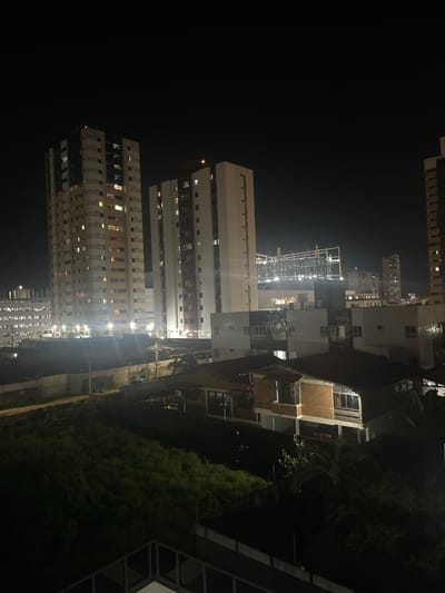 The image depicts a nighttime cityscape in João Pessoa, Brazil, characterized by a collection of buildings of varying heights. Dominating the scene are two tall, modern apartment buildings with numerous illuminated windows, suggesting occupancy. To the left of these, another multi-story structure, possibly a parking garage or a more utilitarian building, is visible. In the mid-ground, a low-rise brick house with a gabled roof and large windows is prominent. Further in the background, more buildings can be seen, including a skeletal framework of a building under construction, indicative of ongoing development. The sky is dark, typical of nighttime, with no visible celestial bodies or aircraft. The foreground is occupied by dark vegetation and the roofline of another building. Streetlights and interior lighting from the buildings illuminate the scene, casting reflections on some surfaces. The overall impression is one of an urban environment at night, with a mix of residential and commercial structures and evidence of construction.