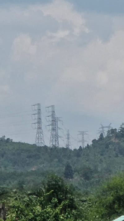 The image shows a landscape with power line towers against a cloudy sky, likely in the rural or semi-rural area near Kon Ra Kiah, Vietnam. The terrain is dominated by green, hilly terrain. The power line towers ascend up the mountain.