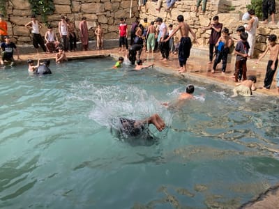 The image displays an outdoor scene at a water recreation area in Killi Malik Sher Mohammad, Pakistan, featuring approximately 25-30 males, predominantly children and adolescents, engaged in activities around and within a man-made pool.
The pool contains light blue-green water. In the immediate foreground, a significant splash of water indicates an individual has just entered, with only their lower legs and feet visible above the surface, along with a partial view of their head/upper body. Several other individuals are in the water, some swimming or standing, including one wearing a dark top on the left and another child in a neon green top towards the center.
Along the concrete edge of the pool, numerous individuals are standing, sitting, or crouching, some appearing to be preparing to enter the water while others observe. Many are shirtless or wear loose-fitting pants or shorts. The background consists of a high, irregularly constructed stone wall made of light-colored rocks. More individuals are visible sitting or standing on top of this stone wall. Green foliage grows on the upper left section of the wall. The scene is brightly lit by natural daylight.