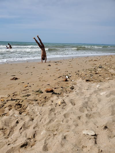 A young person is performing a handstand on a sandy beach with the sea in the background. The person is wearing dark swim trunks. There are waves gently rolling onto the shore. In the distance, two other people are visible in the shallow water. The beach is littered with small stones and some dried seaweed. The sky is partly cloudy, and a distant city skyline can be seen on the horizon.