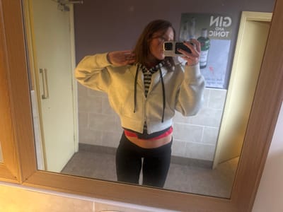 A mirror selfie depicts an individual in what appears to be a public washroom or similar facility in Weymouth, United Kingdom. The subject, a female with brown hair, is positioned centrally, holding a black smartphone with a light-colored case up to her face, obscuring part of it, to capture her reflection. She wears a grey cropped zippered hoodie over a black and white striped top, black leggings, and a visible red band, possibly from an undergarment, at her midriff. Her left hand, with dark nail polish, is raised towards her neck/shoulder area.
The environment includes a large rectangular mirror with a light wooden frame, reflecting the scene. Behind the subject, the wall features light grey square tiles on the lower half and a plain light purple/grey color on the upper half. To the left of the subject's reflection (viewer's right), a light cream-colored door with a horizontal metal handle and a door-closer mechanism at the top is visible. To the right of the subject's reflection (viewer's left), a poster advertising "GIN AND TONIC," featuring a green bottle and text, is affixed to the upper wall. The floor is covered with dark grey square tiles. A light-colored counter or shelf is visible below the mirror frame.
