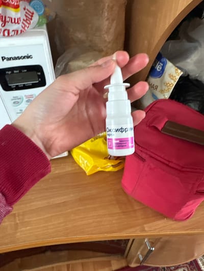 A fair-skinned person's left hand is holding a white nasal spray bottle with a pink label, oriented horizontally. The label has writing in Russian. The person is wearing a red long-sleeved shirt.
The background contains a light-colored wooden surface, a pink bag with a brown handle, a partially visible yellow bag, and a white Panasonic microwave control panel. The location might be Zapadnaya, Russia.