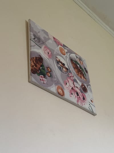 This is a medium shot of a painting of a still life, hanging on a wall. The painting is framed and appears to be a print or a paint-by-numbers canvas. It depicts a table setting with breakfast items and flowers. On the table, there are plates with croissants, blueberries, and what appears to be coffee cups. There are also pink and white flowers arranged on the table, along with a jar with a checkered lid. The overall color palette is soft, with shades of grey, pink, and brown. The painting is hung at an angle on a light-colored wall, with a white molding visible in the top left corner. There is some faint text printed along the bottom edge of the canvas, but it is mostly illegible. The scene is indoors, with no indication of time of day or weather. The painting itself suggests a calm and pleasant breakfast setting.