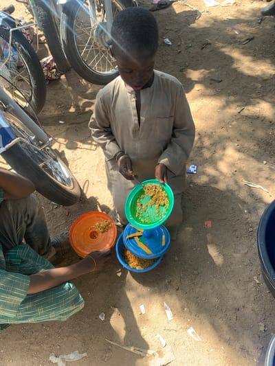 A young boy in Boi A, Nigeria, holds a green plate filled with food. He is wearing a tan tunic. In front of him are three plastic containers, one orange, one blue, and another one with a blue lid. The orange container has food in it. The boy appears to be eating. He is standing on a dirt ground. Two motorcycles are parked behind him. There is also a person sitting next to the boy wearing a green and white checkered shirt.