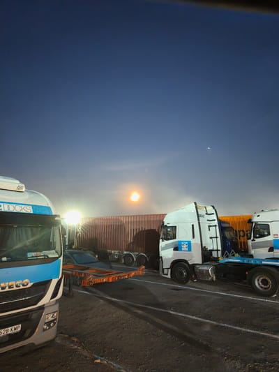 The image shows a truck stop at night in Alcàsser, Spain. Three large white trucks, two of which have "DOCKS" written on their sides, are parked on an asphalt surface marked with white lines. One truck is pulling a flatbed trailer, upon which sits a dark-colored car. Behind the trucks are shipping containers. The sky is dark blue with a bright moon or streetlight visible in the distance. The scene is illuminated by artificial lights, creating long shadows.