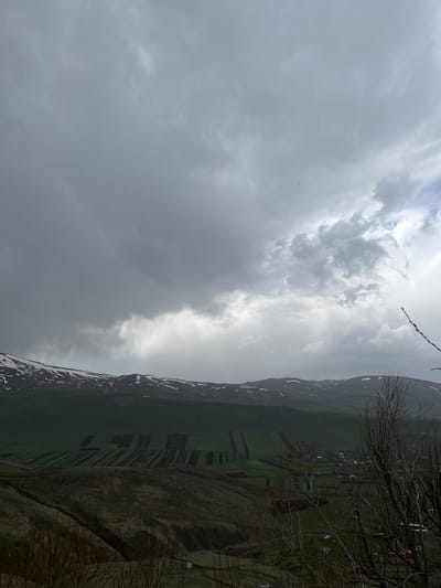 The image depicts a cloudy landscape near Hartagyugh, Armenia. The foreground features barren trees and a descending slope with varied brown and green vegetation. Beyond this, a valley unfolds, displaying a pattern of green fields intersected by dark strips. A mountain range rises in the background, its peaks partially covered in snow. The sky is dominated by heavy, dark clouds, with patches of brighter, diffused light breaking through in places. Scattered structures, likely dwellings or farm buildings, can be discerned in the distance along the edge of the valley.
