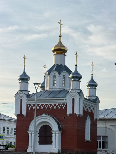 The image shows the Church of the Resurrection of Christ in Cherdyn, Russia. The church is constructed of red brick with white trim and features several onion domes, one large golden dome and four smaller silver domes, each topped with a cross. The architecture appears to be typical of Russian Orthodox churches. In the foreground, there is a streetlamp and part of a sidewalk. To the left of the church, a portion of a white building with multiple windows is visible, along with a yellow pedestrian crossing sign. The sky is overcast with some clouds.