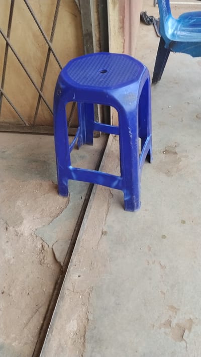The scene depicts a blue plastic stool in an outdoor or semi-enclosed area within Isieke, Nigeria. The stool features an octagonal, textured seat with a central circular hole, supported by four legs connected by horizontal braces. The plastic material shows signs of wear, scuffs, and dirt, with lighter stress marks visible on the leg supports. The stool rests on a worn concrete or cement floor that exhibits cracks, varying shades of gray and beige, and visible dirt. A thin, linear metal strip, likely a track or trim, runs diagonally across the floor, passing underneath the stool. In the immediate background, to the right, a portion of a second blue plastic chair is visible, along with a crumpled dark plastic object on the ground. To the left background, a dark metal grid structure, possibly a gate or security grate, is positioned in front of a light brown wall or panel. The overall environment appears utilitarian and exposed to elements, with no individuals or direct actions observed.