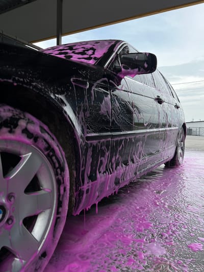 A black sedan, specifically a BMW 3-series model, is shown undergoing a wash at a car wash facility in Velyki Hai, Ukraine. The vehicle is entirely covered in a vibrant pink cleaning foam, which is actively dripping down its black body panels and silver multi-spoke alloy wheels. A BMW logo is discernible on the center cap of the front wheel. The car is situated within a covered bay, indicated by a grey overhead structure and a yellow support beam. Beyond the bay, a cloudy light blue sky is visible, along with distant white industrial-style buildings, a fence, and utility poles. The dark ground surface beneath the car is thoroughly saturated with the pink foam and water runoff, forming distinct puddles and trails of bubbles. No individuals are present in the image.