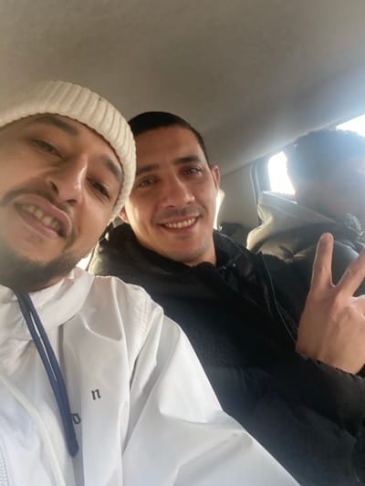The image depicts three male individuals seated inside a vehicle. The specific location within the city of La Wantzenau, France, cannot be determined from the visual information provided.
The individual positioned in the foreground on the left is a male with light complexion, facial hair including a beard and mustache, and is wearing a white knitted beanie and a white jacket with blue drawstrings. He is looking directly at the camera with an open-mouthed smile, his lips slightly pursed in what appears to be a playful or exaggerated expression.
Behind and to the right of the first individual is a second male, also with a light complexion, wearing a dark jacket. He is smiling with teeth visible and looking towards the camera.
Partially visible in the background on the far right is a third male, of darker complexion, wearing a light-colored hooded garment. His right hand is raised, forming a "peace sign" gesture with the index and middle fingers extended.
The environment is the interior of a vehicle, evidenced by the light-colored headliner visible at the top of the frame and a window in the background on the right, through which natural light is entering. The individuals appear to be seated in a row or close proximity, consistent with car seating.