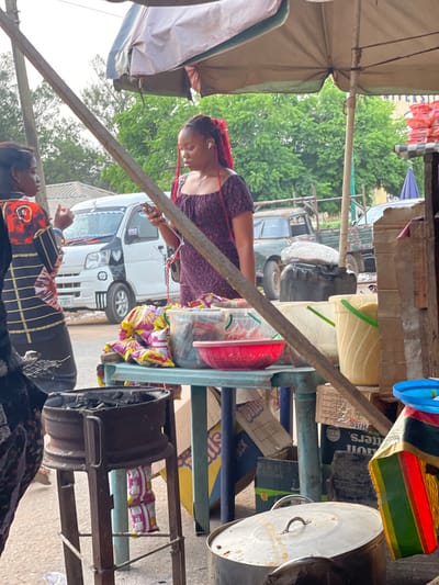 The image captures an outdoor market scene, likely during the daytime, with several people and various goods visible under what appears to be a slightly overcast sky.
In the foreground, a woman with reddish-brown braided hair, possibly dreadlocks or twists, is standing to the left of a makeshift stall. She is wearing a dark, short-sleeved top with a subtle pattern and has white earbuds in her ears. Her gaze is directed downwards, suggesting she might be looking at a phone or an item in her hands.
The stall itself is constructed from metal framing and shelves, laden with various items. On the upper shelves, clear plastic containers hold goods; one contains a red bowl or lid, and another larger container appears to hold light-colored grains or produce. Below these, there are several plastic bags of snacks, identifiable by their pink and white packaging. Cardboard boxes are also stacked on and around the stall, with some partially visible text, including "BUTTERS," "FRUIT," "Dolls" (or "Balls"), and "PACK." A large metal pot with a lid rests on the right side of the stall, and a colorful striped bag (red, yellow, green) hangs near it. A sturdy diagonal beam, possibly part of the stall's support or another nearby structure, crosses the mid-ground.
To the far left, a light-colored fabric canopy or umbrella structure provides shade, supported by visible poles. In the background, several vehicles are parked on what appears to be a road or open area. A white mini-van or small bus with dark decorative accents (possibly heart shapes) on its front is clearly visible behind the woman with red hair. Further back, an older, possibly darker-colored truck or van with a spare tire mounted on the rear is also present. Green foliage from trees is visible above and between the vehicles.
In the bottom right corner, another woman is partially visible, wearing a dark top with orange and black patterns. She appears to be looking upwards or towards the left, possibly interacting with someone or observing the scene.
The overall atmosphere suggests a busy, utilitarian market environment.