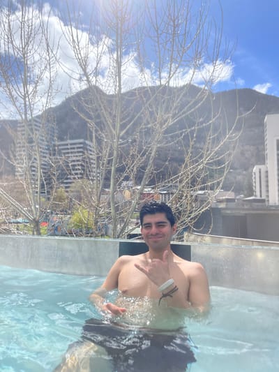 A person, identified as male, is chest-deep in a pool of water outdoors. The person is shirtless, facing toward the camera, and giving a hand gesture. The body of water appears to be an outdoor hot tub or pool, as the surroundings indicate an urban area. In the background, the city of Les Escaldes, Andorra, is visible, including buildings and a mountain range. In the foreground are a bare tree and water droplets.