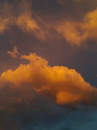 The image displays a dramatic sky filled with clouds at sunset. The prominent clouds are illuminated by the setting sun, giving them a vibrant orange hue that contrasts with the darker, moody tones of the surrounding sky. The clouds are thick and billowy, with some appearing more wispy and ethereal. The lighting suggests a transition from day to night, with warm golden light hitting the clouds from the side. The overall mood is serene yet powerful, capturing the beauty and transient nature of a sunset. No people, objects, or text are visible. The scene is set outdoors, likely in a location with an open sky, as indicated by the vast cloud formations. The context provided places the image in Tash-Dobo, Kyrgyzstan.