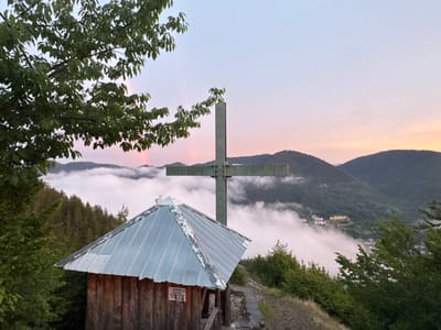 This media file captures a serene mountain landscape at dusk, with a prominent metallic cross and a small wooden structure in the foreground. The setting appears to be a hilltop in Strazha, Bulgaria, indicated by the location context provided.
In the foreground, a rustic wooden hut with a corrugated metal roof is positioned to the left. Attached to the side of the hut is a red and white sign with text in Cyrillic script. Behind the hut, a large, dark metallic cross stands tall, dominating the mid-ground.
The background reveals a breathtaking view of rolling mountains partially obscured by a thick layer of white clouds or fog, which creates a mystical atmosphere. A faint rainbow can be seen arching through the sky above the fog. In the distance, nestled among the lower slopes of the mountains, are buildings that suggest a small settlement or resort, adding a touch of human presence to the otherwise natural panorama.
The time of day is clearly evening, as indicated by the soft, pastel hues of the sky – a blend of pale blue, pink, and orange, typical of a sunset. The weather appears calm, with the fog contributing to a sense of peacefulness. The overall emotion conveyed is one of tranquility and spiritual reflection, amplified by the presence of the cross and the natural beauty of the scene. The lush green trees framing the image further enhance the sense of being in nature.
The visible text on the sign on the wooden hut appears to be in Bulgarian and reads: "ЗАБРАНЕНО ЗА МИНИСТЕРСКИТЕ КУРАЛИ". This translates to "Forbidden for Ministerial Hangers-on" or "Forbidden for Ministerial hangers-on".