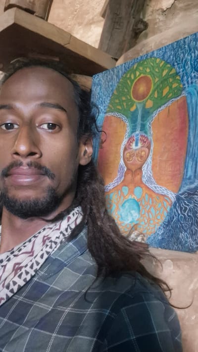 A close-up, selfie-style shot captures a person in the foreground, with a painting visible to their right. The person, with dark skin, dark eyes, and dark, dreadlocked hair pulled back, is wearing a patterned scarf and a green and blue plaid shirt. They have a goatee and mustache. The painting depicts a stylized, abstract human form with a tree-like structure emanating from the head, set against a blue, watery background. The scene appears to be indoors, with wooden beams and rough-textured walls suggesting a rustic or natural environment. The lighting is soft and natural. No specific location within the city of Shila, India, can be identified from the image.