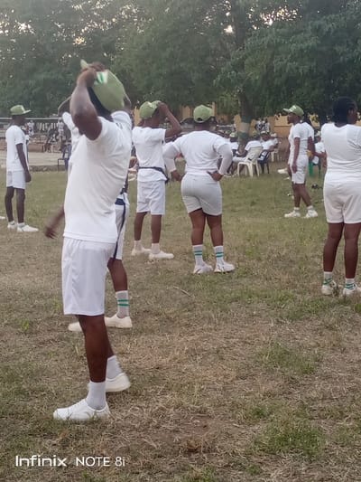 A group of people wearing white t-shirts, shorts, white sneakers, and green hats stand on a grassy field. Some are adjusting their hats. In the background, other people are seated in chairs. A building and trees are also visible in the background.