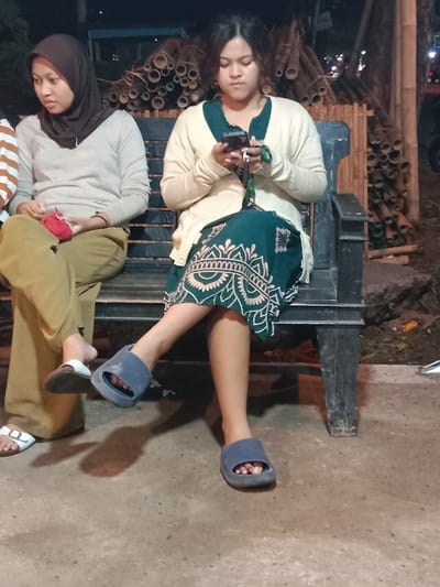 Two young adult females are seated on a dark-colored bench outdoors at night in Sekopek Wetan, Indonesia. The female on the left, wearing a dark brown hijab, a light-colored long-sleeved top, and olive pants, is holding a small red object and looking to her left. She wears white sandals. The female on the right, with dark hair, is wearing a cream-colored cardigan over a dark green patterned dress/skirt and blue slip-on sandals. She is actively engaged with a black smartphone, holding it with both hands and looking down at the screen. Her legs are crossed at the knees. In the background, a substantial stack of cut bamboo poles is visible. The ground surface is light-colored concrete or pavement. The scene is illuminated by ambient artificial light sources, indicating nighttime conditions.