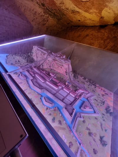 The image shows a detailed scale model of a fortress within a glass display case. The model represents a bastioned fort design with water-filled moats surrounding fortified structures, including buildings and ramparts. The layout includes a town section integrated with the defensive architecture. The environment outside the fort is modeled with trees and landscape features. The model is likely located inside a historical site or museum, based on the surrounding environment of a cave or stone structure with a rough texture. The location is within the city of Homburg, Germany.