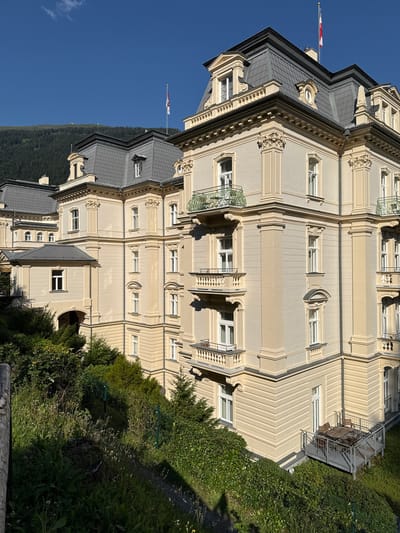 The image shows the Kaiserhof Hotel in Bad Gastein, Austria, a grand Belle Époque building with a mansard roof and ornate detailing. The hotel is situated on a hillside, with lush green vegetation and trees in the foreground and a mountainous, forested landscape in the background under a clear blue sky. The hotel's facade is painted a pale yellow, with cream-colored trim and decorative elements. Several balconies with wrought-iron railings are visible, and one on the right side has outdoor furniture. The hotel has a prominent mansard roof with dormer windows and a flagpole flying a red and white flag. The architecture suggests a historic and elegant establishment.