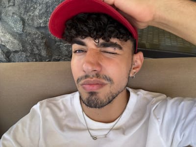 A young male individual is depicted in a close-up, self-portrait style photograph. He has curly dark hair, light brown skin, and a trimmed beard and mustache. He wears a red baseball cap, which his right hand is partially grasping or adjusting at the top of the frame. His left eye is open, while his right eye is squinted, creating a winking expression, and his lips are slightly pursed. He has two silver stud piercings on his left eyebrow and multiple silver piercings on his left ear, including studs on the helix/cartilage and a hoop on the lobe. He is dressed in a plain white crew-neck t-shirt and a silver chain necklace with a safety pin pendant featuring a small, ornate, skull-like detail. The immediate background consists of a light brown upholstered surface. Behind this surface, a grey, rough-hewn stone wall is visible on the left, and a dark, reflective surface, possibly a window, is on the right. The specific location within the city of Ia Municipal Unit, Greece, cannot be determined from the visual information provided.