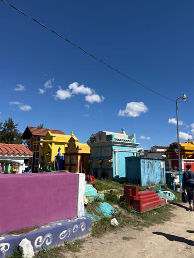 A vibrant cemetery under a bright blue sky with scattered clouds is depicted. Colorful tombs and mausoleums in hues of yellow, blue, purple, and red are arranged along a path. A utility pole with a speaker stands tall, wires stretching across the sky. Grass grows beside the pathway, casting shadows. A person walks along the edge of the cemetery.