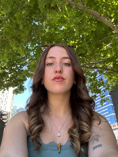 A selfie showcases a young woman with brown hair styled in loose waves, wearing a green top and two delicate necklaces. She has fair skin, green eyes, and a nose piercing. Sunglasses are perched atop her head. A tattoo is visible on her upper arm. The background features lush green trees and the tops of modern city buildings under a bright sky. The perspective is slightly angled upwards.