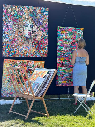This image captures an outdoor art exhibition, likely in a village setting, as suggested by the presence of a tent structure and lush green grass. The foreground is dominated by a wooden display rack filled with various framed artworks, suggesting a focus on visual arts. In the midground, three large, vibrant paintings are displayed against a black backdrop. The leftmost painting is a detailed mosaic-like portrait of a woman surrounded by colorful butterflies and intricate patterns. To its right is a tall, narrow painting composed of numerous repetitions of the word "love" in various fonts and colors. Below these, a horizontally oriented painting depicts a floral arrangement with a yellow background. A woman, dressed in a denim sundress and red flats, is visible in the right portion of the image, observing the artwork. Her stance and focus indicate engagement with the art. The environment appears to be a sunny day, with dappled light on the grass, and a white folding chair is positioned to the right, adding to the outdoor exhibition feel. The overall scene suggests a community event or art fair, possibly within the Village of Sag Harbor, United States of America, given the mention of a village context.