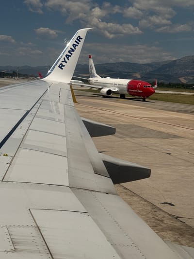 Two airplanes are on a tarmac under a partly cloudy sky. In the foreground, the wing of a Ryanair airplane is visible, with "RYANAIR" written in blue on the white wingtip. In the middle ground, a Norwegian Air Shuttle airplane, with a red nose and "norwegian.com" written on the side, is taxiing on the runway. In the background, mountains and trees are visible. The scene takes place at Tirana International Airport, which is located in Kruja, Albania.