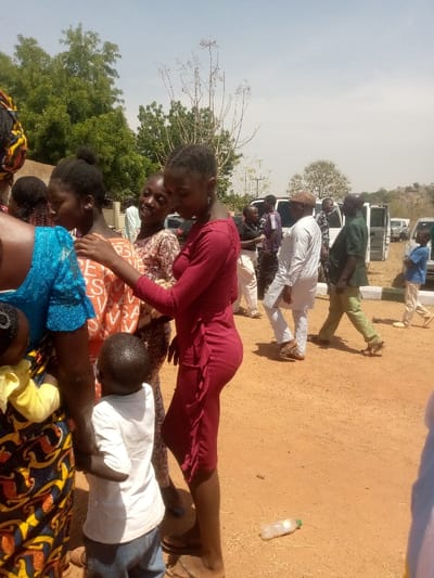 The image displays an outdoor scene in Kagoma, Nigeria, characterized by a reddish-brown dirt ground under a clear, bright sky. In the immediate foreground, a small child, estimated to be between 2-4 years old, is visible from the rear, wearing a white t-shirt and blue jeans. This child is positioned between a partially visible woman in a blue patterned top on the left and a young woman in a form-fitting, long-sleeved, ruffled red dress, standing centrally and looking downwards. To the left of the red-dressed woman, another woman in an orange patterned top smiles while placing her right hand on the red-dressed woman's right shoulder.
In the midground and background, approximately a dozen adults and several children are observed standing and walking. Notable individuals include a man in white traditional attire and a brown cap, and a man in a dark green shirt and light green pants, both appearing to walk towards the right side of the frame. Several light-colored vehicles, consistent with vans, are parked in the background. A clear plastic water bottle lies horizontally on the dirt ground in the lower right foreground. The background features green trees and sparse, dry vegetation, with low hills visible on the far right horizon.