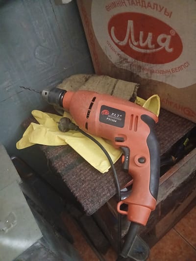The image displays a close-up view of an orange and black power drill resting on a brown, textured surface, possibly a piece of furniture or fabric. A pair of bright yellow gloves is positioned beneath the drill. In the background, a large bag with text in Cyrillic script and a red oval logo is visible. The text includes the word "Лия" within the logo. The environment appears to be indoors, with a somewhat cluttered and utilitarian aesthetic, suggesting a workshop or storage area. The overall lighting is dim, with some areas being more illuminated than others, highlighting the drill and gloves. The exact location of Shakhtinsk, Kazakhstan, is not depicted in the image itself, but the context provided suggests a possible setting.