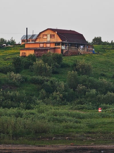 A wide shot captures a cluster of wooden buildings situated on a grassy hill in Potapovo, Russia. The primary structure is a large, two-story house with a dark red, sloping roof and wooden siding. It features multiple windows and a covered porch with a railing. In front of the house, a smaller, attached wooden structure appears to be a shed or workshop. To the left, another building with a lighter blue roof and a tall, dark chimney is visible. The surrounding landscape is characterized by rolling green hills covered in lush vegetation, with scattered trees and shrubs. In the foreground, the terrain transitions to darker, muddier ground. A red and white cylindrical marker stands on the grassy slope to the right of the frame. The sky above is overcast with a light gray hue, indicating diffused daylight. There are no people visible in the scene.