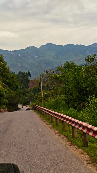 The image shows a road in Minh Tân Commune, Vietnam. The road curves to the left and leads downhill. On the right side of the road is a guardrail with alternating red and white stripes. Lush green vegetation grows on either side of the road, with taller trees and shrubs forming a dense backdrop. In the distance, rolling hills and mountains are visible under a cloudy sky. A utility pole stands along the roadside, with power lines stretching across the scene. A vehicle, possibly a motorcycle, is seen farther down the road.