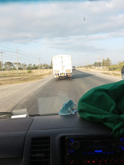 A view from inside a vehicle shows a long, straight road in a rural area. A white truck travels ahead, with another vehicle visible further down the road. Power lines run along the side of the road. Patches of dry vegetation and trees are scattered across the landscape. The dashboard of the vehicle is visible, with a green cloth and a light blue object resting on it.
