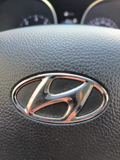 The image shows a close-up of a Hyundai logo on a steering wheel, likely inside a car. The steering wheel appears to be made of black leather with a textured surface. The Hyundai logo is silver and oval-shaped, with the distinctive stylized 'H' inside. In the background, out of focus, are parts of a car's dashboard, including illuminated gauges with purple lights. The lighting suggests it is daytime, and the car is stationary. There are no people visible in the image. The image does not provide any information about the location in the city of Kladno, Czechia.