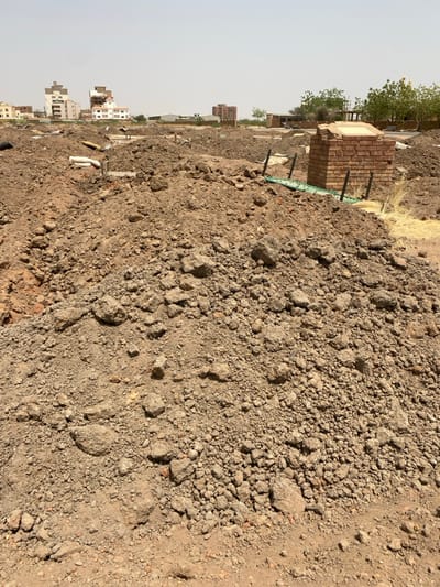 The image shows a pile of dirt, likely a dump site, located in Khartoum, Sudan at latitude: 15.528, longitude: 32.571.  A brick structure partially buried in the dirt can be seen in the distance. The dirt is loose, brown and appears to be dry. Buildings and trees are visible in the background, indicating a suburban area.  The sky is clear and bright.  No people are present.