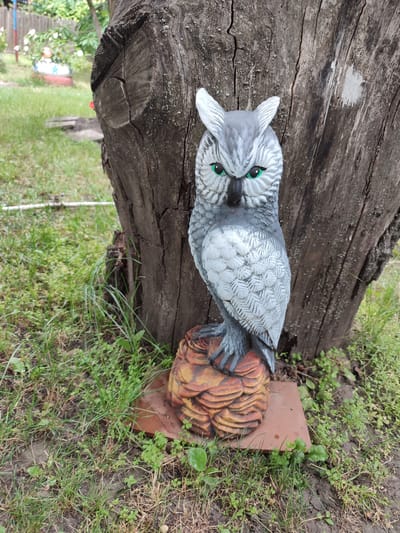 A garden statue of a grey owl with bright green eyes stands on a textured brown base in front of a large, rough-barked tree. The owl is positioned to the left of the tree trunk. The ground around the tree is covered in green grass and some scattered brown leaves. In the background, a wooden fence and greenery are visible. The scene appears to be in an outdoor setting, likely a garden or yard. This image was taken in Voskobiinyky, Ukraine.