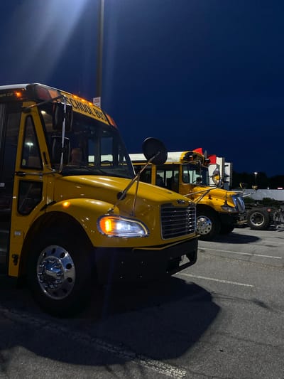 Two yellow school buses are parked in a lot at dusk. The bus in the foreground is facing left, while the bus behind it is facing right. The sky is dark blue, and there is a light pole in the background. The ground is asphalt with painted parking lines. A person is visible in the driver's seat of the foreground bus.