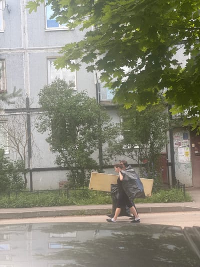 Two individuals are walking on a sidewalk in front of a tiled apartment building in Zhilgorodok, Russia. The person in front, a female, is carrying a large, flat, light-colored object, possibly a piece of furniture or construction material, under her left arm and shoulder. She is wearing a dark dress and white socks. Behind her, a second individual, possibly male, is carrying a large black trash bag, which is draped over their left shoulder and arm, and partially covering the object being carried by the first person. This individual is wearing dark clothing and dark shoes. The apartment building has a grey, patterned tiled facade with numerous windows. Green trees and foliage are visible in front of and around the building. A grey curb and sidewalk separate the building from a dark grey surface in the foreground, which appears to be a wet road or a reflective surface like a puddle or a car roof, reflecting the building and trees. The lighting suggests it is daytime.