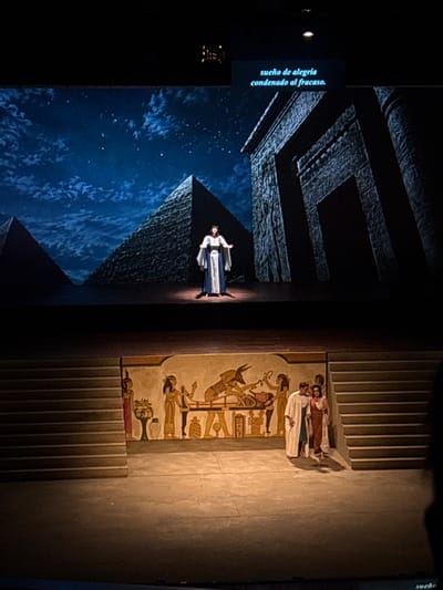 The image captures a theatrical scene in San Borja, Peru. A performer in ancient Egyptian garb stands on a set resembling a pyramid against a backdrop of a starry night. Nearby, a wall is adorned with Egyptian-style drawings. To the right of the wall, a set of stairs lead upwards, where a small group of people can be seen. To the far left, there's a black screen with the words "sueño de alegria condenado al fracaso," Spanish for "dream of joy condemned to failure." The overall lighting suggests a stage production, with focused illumination on the performer and the set elements.