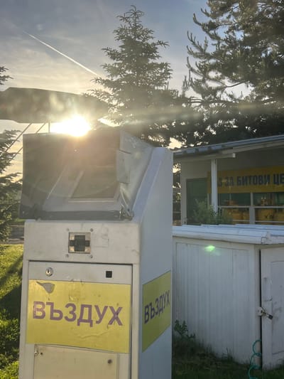 The image shows a view of a structure in Dragushinovo, Bulgaria, with a sunflare obscuring parts of the scene. In the foreground, a beige machine with a yellow sign bearing the Cyrillic text "ВЪЗДУХ" (Vazduh, meaning "Air") is visible. To the right, a white building with a metal roof and a yellow sign with Cyrillic text is partially visible. Behind the structures, pine trees are silhouetted against the sky, which features a contrail from an aircraft. The lighting suggests it is either sunrise or sunset.